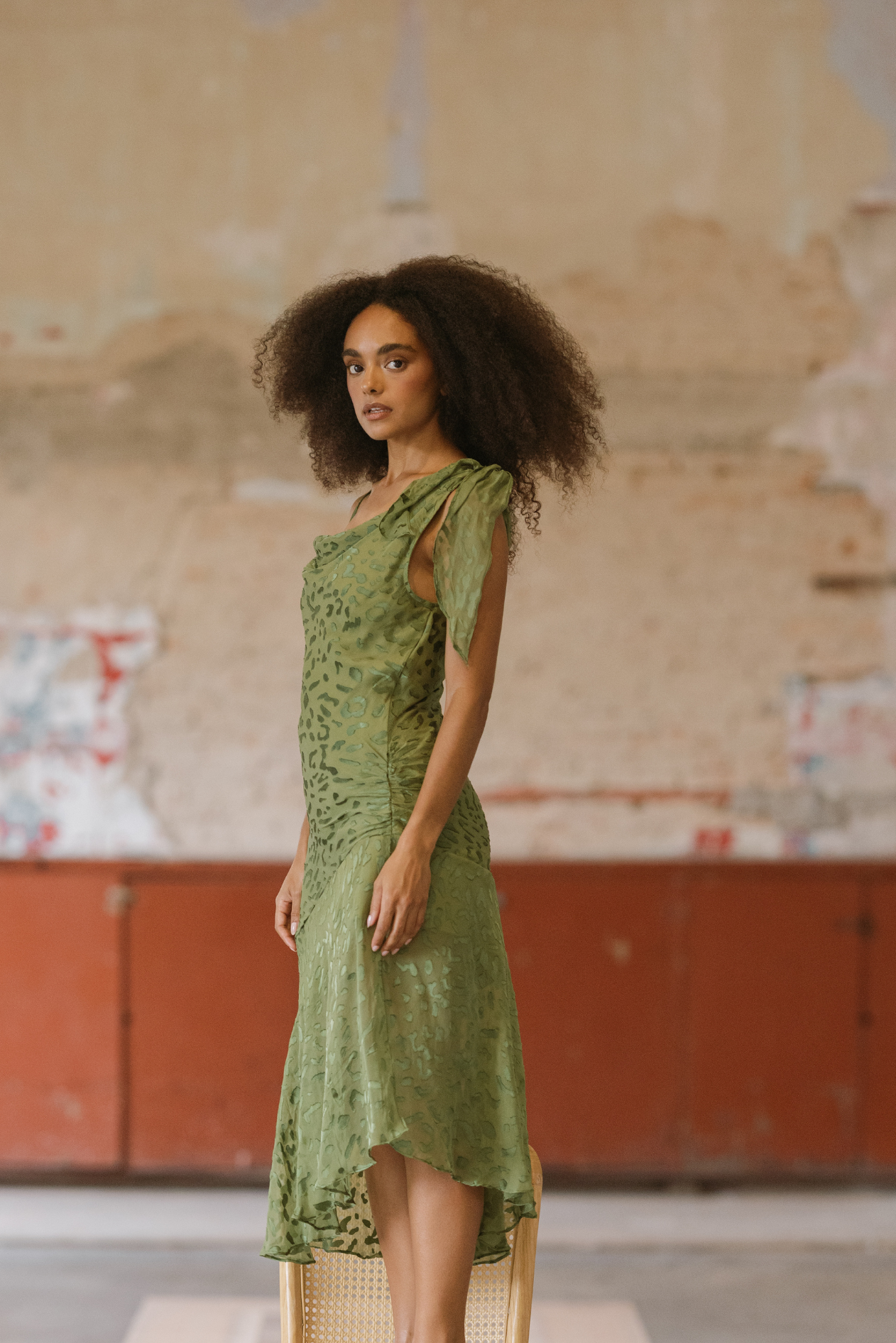 Woman in a green dress sitting on a chair against a textured wall.
